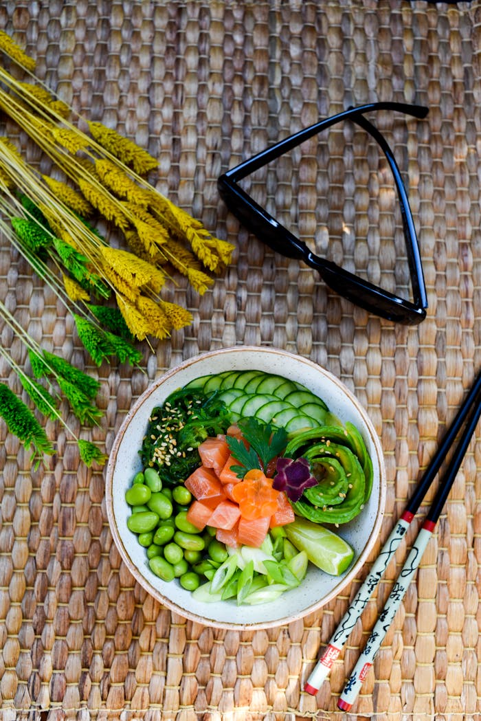 Colorful sushi bowl with fresh vegetables, chopsticks, and sunglasses on a woven mat. Perfect for healthy food concepts.
