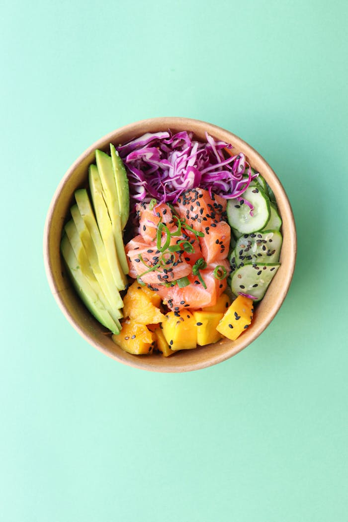 Top view of healthy poke bowl with fresh blue cabbage with chopped avocado cucumber and mango with raw salmon on green background