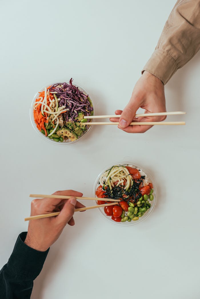 Two people using chopsticks to enjoy fresh vegetable bowls on a white table.